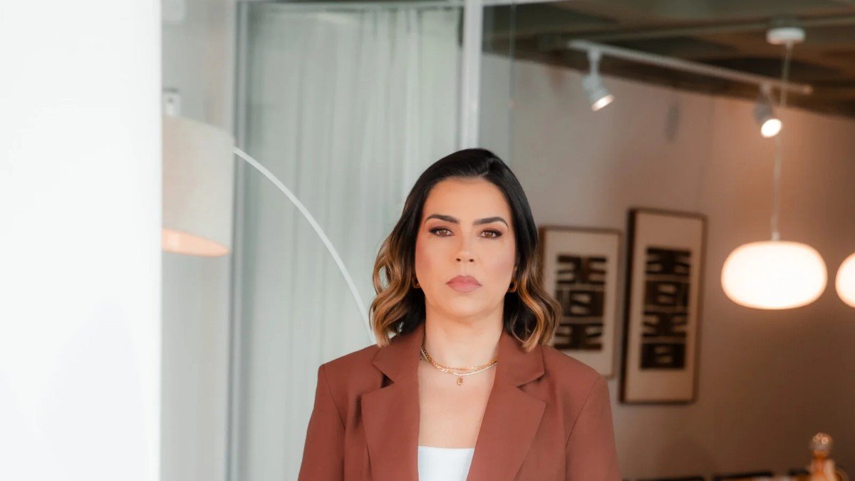 Luciana Prates, de blazer marrom, em um escritório com quadros ao fundo