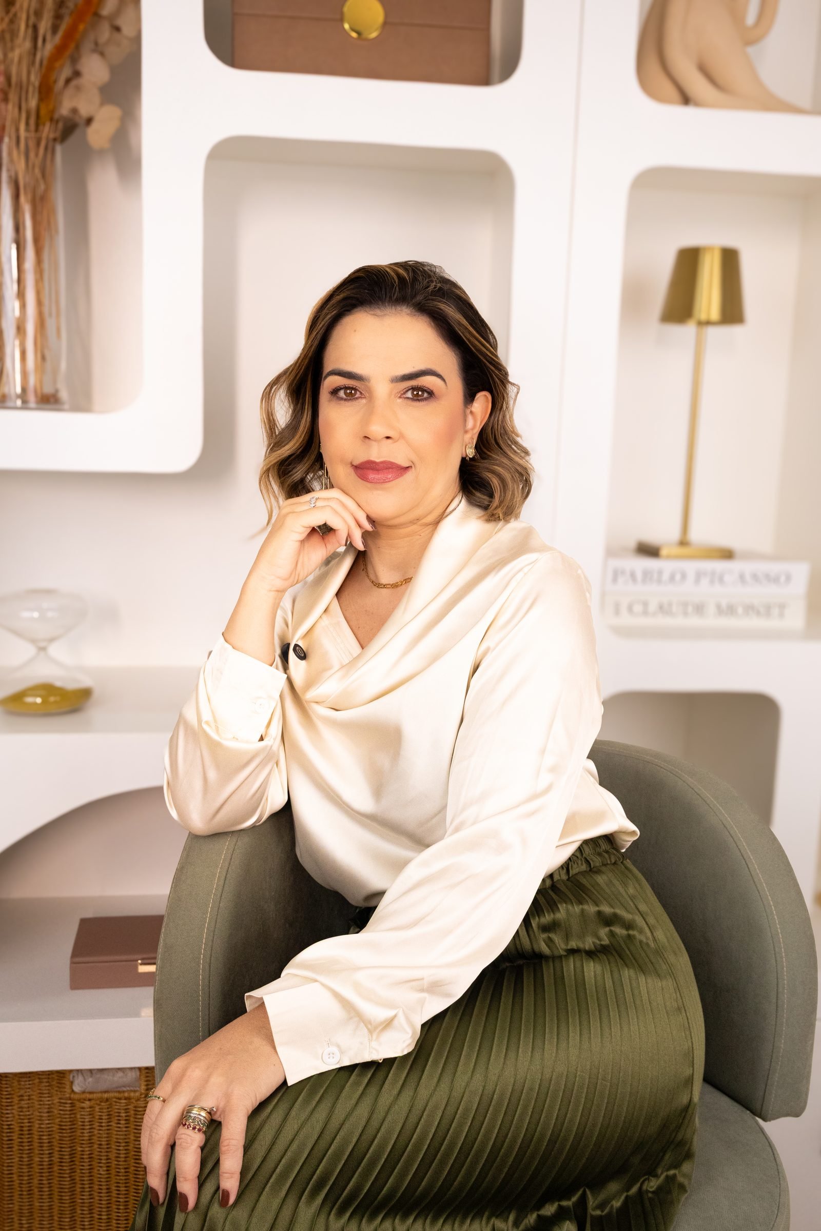 Luciana Prates, de blusa de cetim clara, sentada em uma poltrona verde em um escritório claro com estante de nichos.