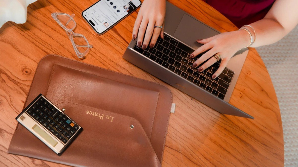Mãos digitando em um notebook sobre uma mesa de madeira, ao lado de um celular e uma agenda